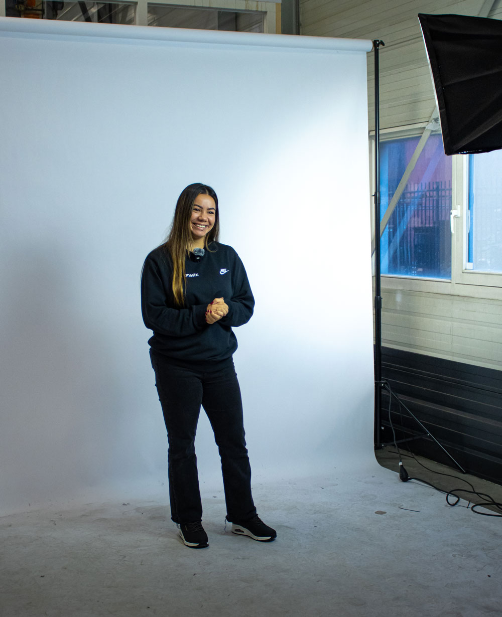 Smiling Jennifer (Jennifer of Onesix) standing in front of a white photography backdrop in a studio with lighting equipment and a window in the background.